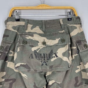 K&ouml;nnte beinhalten: Tarnfarbene Cargohose mit mehreren Taschen und Knopfverschl&uuml;ssen. Die Hose hat das Logo "AVIREX" auf der Vordertasche aufgedruckt. Die Farbpalette umfasst Gr&uuml;n-, Braun- und Beiget&ouml;ne.