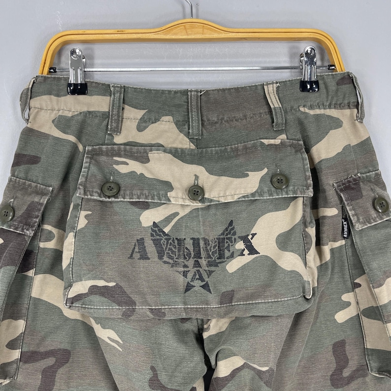 K&ouml;nnte beinhalten: Tarnfarbene Cargohose mit mehreren Taschen und Knopfverschl&uuml;ssen. Die Hose hat das Logo "AVIREX" auf der Vordertasche aufgedruckt. Die Farbpalette umfasst Gr&uuml;n-, Braun- und Beiget&ouml;ne.