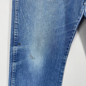 Pode incluir: Close-up de uma se&ccedil;&atilde;o de jeans azuis. Os jeans mostram sinais de desgaste e desbotamento, com algumas pequenas manchas. As costuras s&atilde;o vis&iacute;veis ao longo das costuras, e o tecido tem uma apar&ecirc;ncia ligeiramente texturizada.