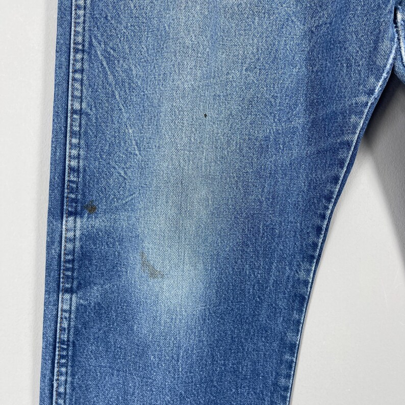 Pode incluir: Close-up de uma se&ccedil;&atilde;o de jeans azuis. Os jeans mostram sinais de desgaste e desbotamento, com algumas pequenas manchas. As costuras s&atilde;o vis&iacute;veis ao longo das costuras, e o tecido tem uma apar&ecirc;ncia ligeiramente texturizada.