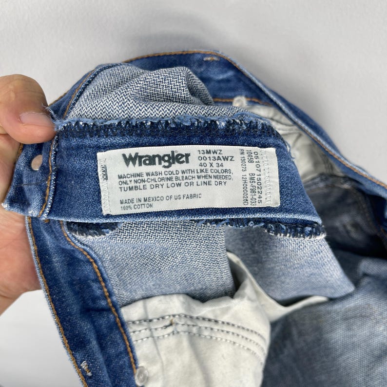 Pode incluir: Close-up do interior de um par de jeans Wrangler azuis. A etiqueta dos jeans &eacute; vis&iacute;vel, exibindo a marca, o tamanho 40 x 34 e as instru&ccedil;&otilde;es de cuidado. A etiqueta tamb&eacute;m informa que os jeans s&atilde;o feitos no M&eacute;xico com tecido dos EUA, 100% algod&atilde;o.