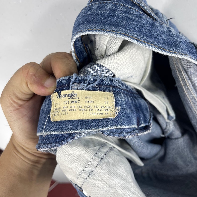 May include: A close-up of a blue denim Wrangler brand jean tag with the style number 0013MWZ, waist size 34, and length 32. The tag has instructions for washing and drying the jeans.