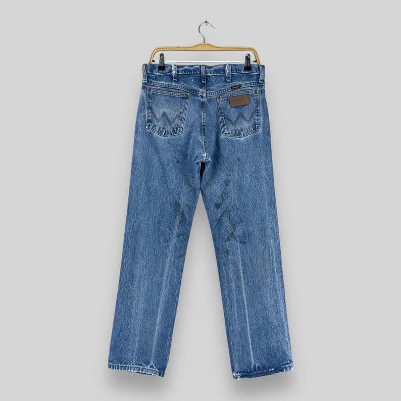 Pu&ograve; includere: Un paio di jeans blu in denim con una toppa in pelle marrone sulla tasca posteriore che dice "Wrangler".
