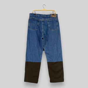 K&ouml;nnte beinhalten: Blaue Jeans aus Denim mit braunen Canvas-Patches an den Knien. Die Jeans h&auml;ngen an einem goldenen Kleiderb&uuml;gel.