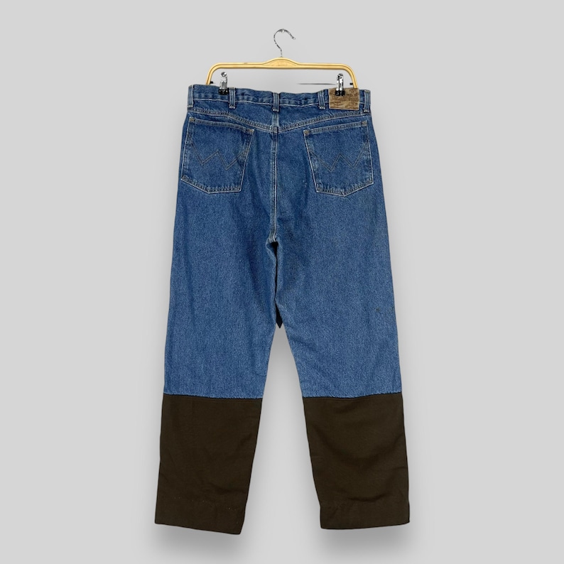 K&ouml;nnte beinhalten: Blaue Jeans aus Denim mit braunen Canvas-Patches an den Knien. Die Jeans h&auml;ngen an einem goldenen Kleiderb&uuml;gel.
