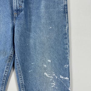 以下が含まれることがあります： 白いペンキの飛沫が付いた、明るい青色のデニムジーンズのクローズアップ。ジーンズは、クラシックな5ポケットのデザインとボタンとジッパーの留め具が付いています。デニムは少し色あせた外観をしています。
