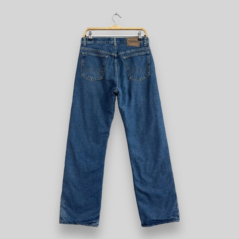 May include: A pair of blue denim jeans hanging on a wooden hanger. The jeans have a brown leather patch on the back with the word "Wrangler" printed on it.