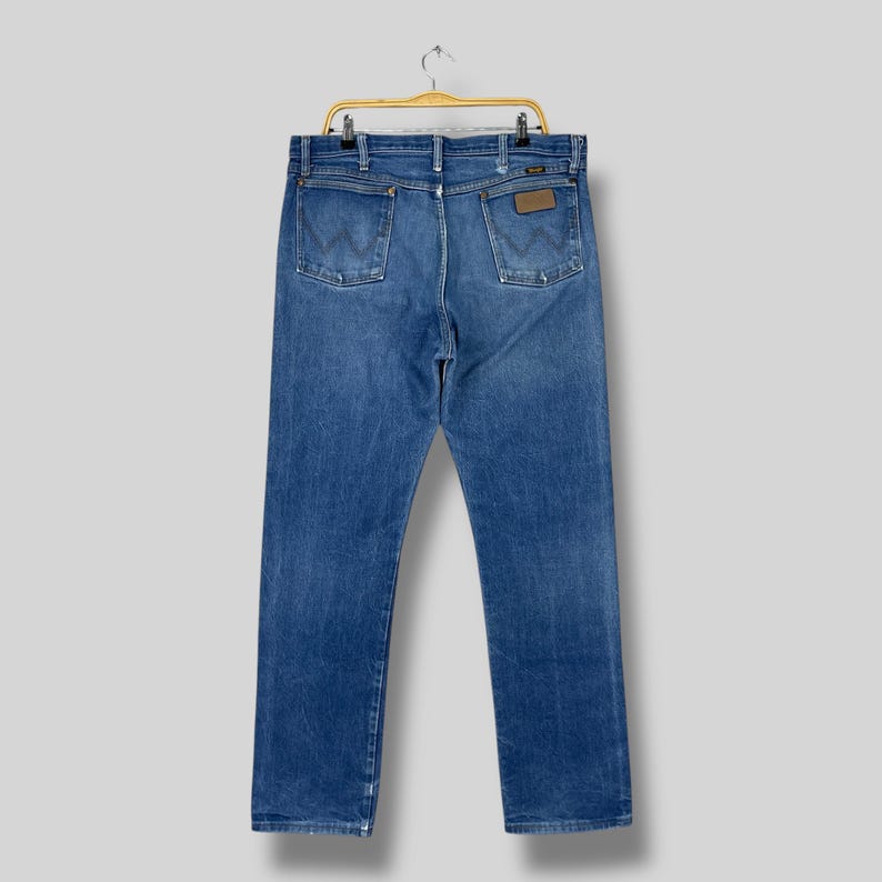 Pode incluir: Cal&ccedil;as de ganga azuis penduradas num cabide dourado. As cal&ccedil;as de ganga t&ecirc;m um design cl&aacute;ssico de cinco bolsos, com o emblema do log&oacute;tipo Wrangler no bolso de tr&aacute;s. As cal&ccedil;as de ganga s&atilde;o de lavagem m&eacute;dia com algum desbotamento.