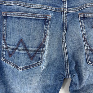 May include: Close-up of the back of blue denim jeans. The jeans feature two back pockets, with the left pocket showcasing a distinctive stitched "W" design in a contrasting brown thread. The denim has a worn, faded appearance.