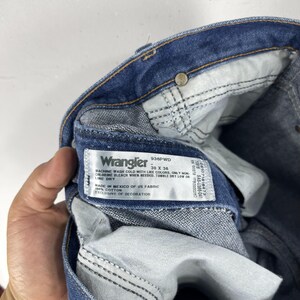 K&ouml;nnte beinhalten: Nahaufnahme eines blauen Wrangler-Jeans-Etiketts mit dem Text "Wrangler 936PWD - 30 X 36" und Pflegehinweisen. Das Etikett ist an der Innenseite der Jeans befestigt.