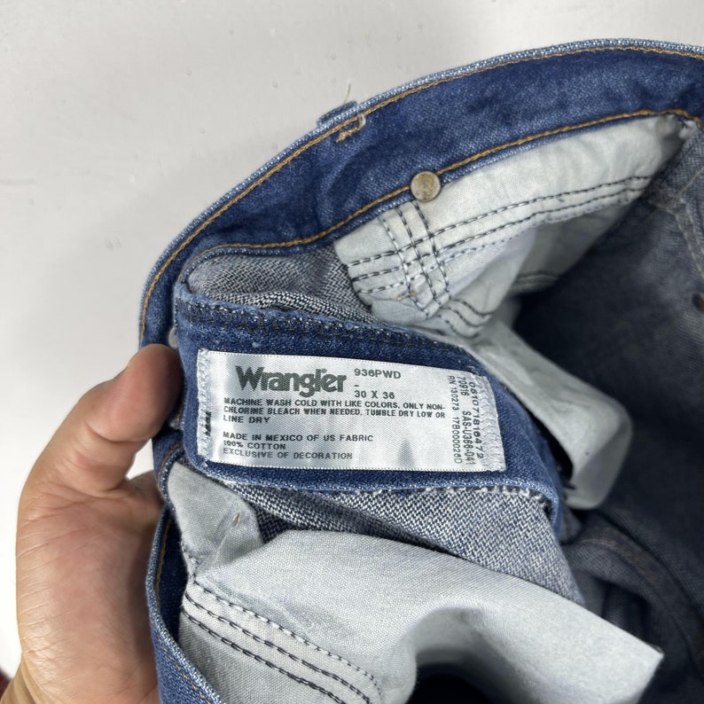 K&ouml;nnte beinhalten: Nahaufnahme eines blauen Wrangler-Jeans-Etiketts mit dem Text "Wrangler 936PWD - 30 X 36" und Pflegehinweisen. Das Etikett ist an der Innenseite der Jeans befestigt.