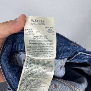 May include: Close-up of a blue denim jeans tag with text including "W33 L34 GREENSBORO" and "MADE IN PAKISTAN." The tag also features washing instructions and size information.