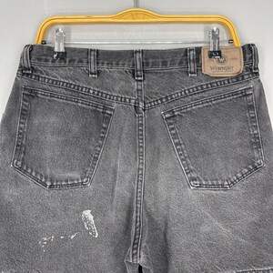 Pode incluir: Close-up da parte de tr&aacute;s de um par de cal&ccedil;&otilde;es de ganga cinzentos Wrangler. Os cal&ccedil;&otilde;es t&ecirc;m dois bolsos traseiros, presilhas para cinto e um patch da marca Wrangler. H&aacute; uma mancha de tinta branca na perna esquerda.