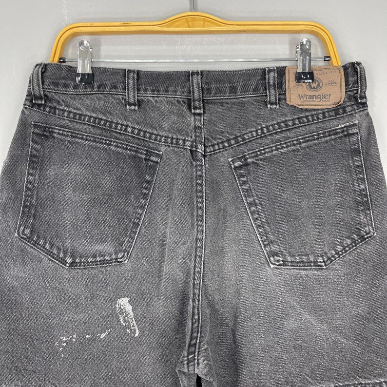 Pode incluir: Close-up da parte de tr&aacute;s de um par de cal&ccedil;&otilde;es de ganga cinzentos Wrangler. Os cal&ccedil;&otilde;es t&ecirc;m dois bolsos traseiros, presilhas para cinto e um patch da marca Wrangler. H&aacute; uma mancha de tinta branca na perna esquerda.