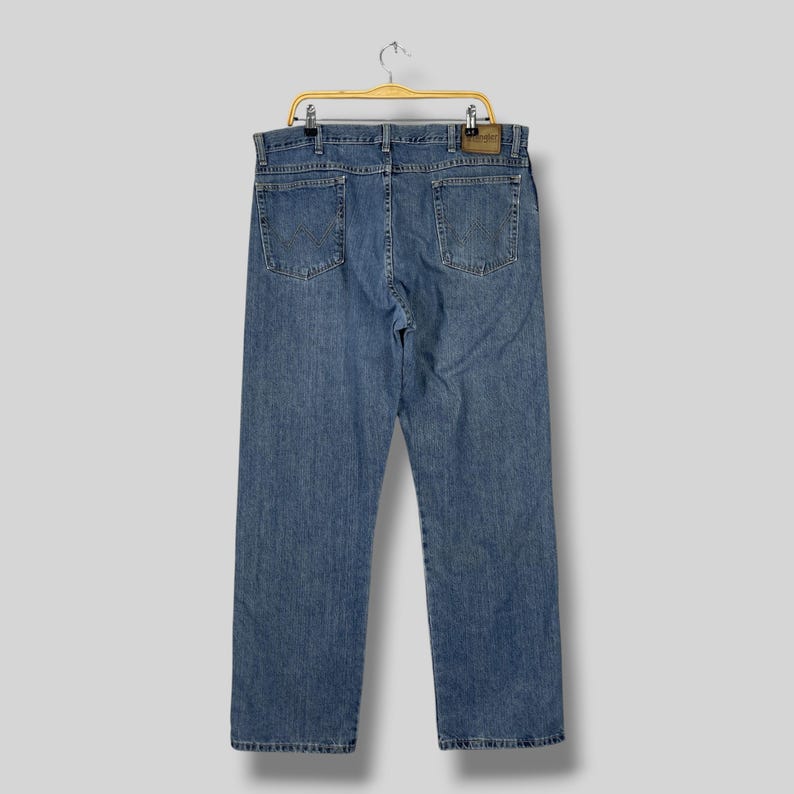 May include: Light blue denim jeans hanging on a wooden hanger. The jeans have a classic five-pocket design, with a leather patch on the back. The jeans are a medium wash and appear to be in good condition.
