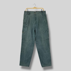 May include: Teal cargo trousers hanging on a gold hanger. The trousers feature multiple pockets on the sides and front, with a button and zip fastening. The fabric appears to be a soft, worn material.