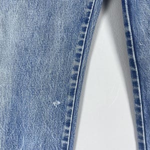 Pu&ograve; includere: Primo piano di un paio di jeans in denim blu chiaro con un lavaggio sbiadito e un aspetto leggermente rovinato. I jeans hanno una gamba dritta e un design classico a cinque tasche.