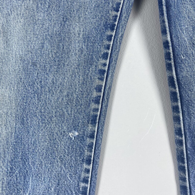 Pu&ograve; includere: Primo piano di un paio di jeans in denim blu chiaro con un lavaggio sbiadito e un aspetto leggermente rovinato. I jeans hanno una gamba dritta e un design classico a cinque tasche.