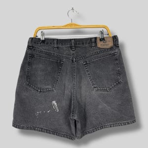 Pode incluir: Cal&ccedil;&otilde;es Wrangler de ganga preta pendurados num cabide amarelo. Os cal&ccedil;&otilde;es t&ecirc;m um design cl&aacute;ssico de cinco bolsos, uma etiqueta Wrangler e um aspeto desbotado e usado. Os cal&ccedil;&otilde;es s&atilde;o de cor cinzenta escura e mostram sinais de desgaste.