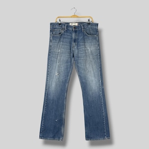 Op de afbeelding: Een medium blauwe spijkerbroek van denim met bootcut pijpen, een klassiek ontwerp met vijf zakken, een knoop- en ritssluiting, en subtiele slijtage- en vervagingsdetails op de voorzijde, hangend aan een goudkleurige metalen kledinghanger.