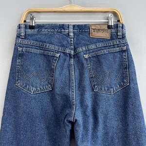 May include: A pair of blue denim jeans with a brown leather Wrangler label on the back pocket. The jeans have a classic five-pocket style and a straight leg.