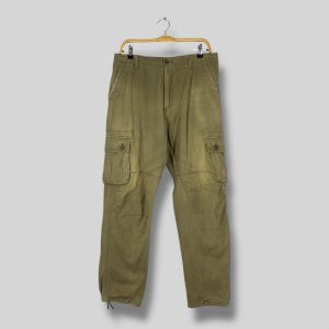 Könnte beinhalten: Olivgrüne Cargohose mit mehreren Taschen und einem Knopfverschluss. Die Hose hängt an einem Holzbügel vor einem hellgrauen Hintergrund. Die Hose hat eine lockere Passform und ein verwaschenes Aussehen.
