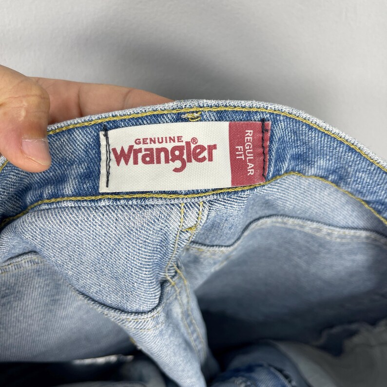 Puede incluir: Primer plano de unos vaqueros de mezclilla azul claro con una etiqueta "Genuine Wrangler". La etiqueta es blanca con texto rojo y una etiqueta rectangular roja que dice "Regular Fit". Los vaqueros est&aacute;n doblados.