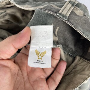 K&ouml;nnte beinhalten: Nahaufnahme eines Stoffes mit Tarnmuster und einem Etikett mit dem Logo "Avirex AUTHENTIC PRODUCT". Der Stoff hat Farbt&ouml;ne von Gr&uuml;n, Beige und Grau. Das Etikett enth&auml;lt Text auf Japanisch.