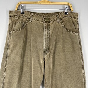 May include: A pair of khaki-colored denim jeans, hanging on a metal and wood hanger. The jeans have a classic five-pocket design, with visible stitching and a button and zipper closure. The jeans are a neutral color.