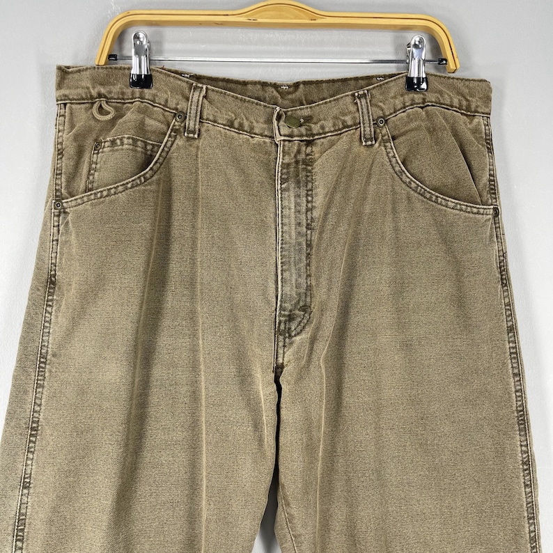 May include: A pair of khaki-colored denim jeans, hanging on a metal and wood hanger. The jeans have a classic five-pocket design, with visible stitching and a button and zipper closure. The jeans are a neutral color.