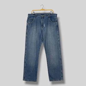May include: Light blue denim jeans hanging on a gold hanger. The jeans have a classic five-pocket design, a button and zipper closure, and a faded wash. The jeans are a relaxed fit.