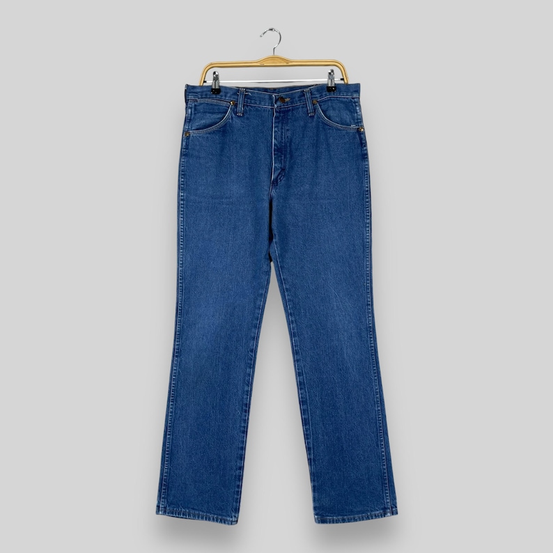 May include: A pair of blue denim jeans hanging on a wooden hanger. The jeans have a classic straight leg style and a button fly.