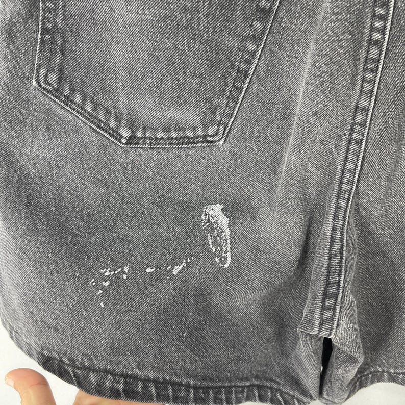 Pode incluir: Close-up de cal&ccedil;&otilde;es de ganga cinzentos com um bolso traseiro e salpicos de tinta branca. Os cal&ccedil;&otilde;es t&ecirc;m um design cl&aacute;ssico com costuras vis&iacute;veis e fecho de bot&atilde;o. O tecido parece gasto e desbotado.