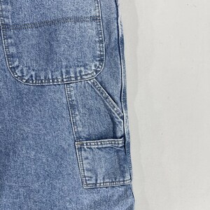 K&ouml;nnte beinhalten: Nahaufnahme einer blauen Jeans-Latzhose mit mehreren Taschen. Die Taschen sind mit einem kontrastierenden dunklen Faden vern&auml;ht.