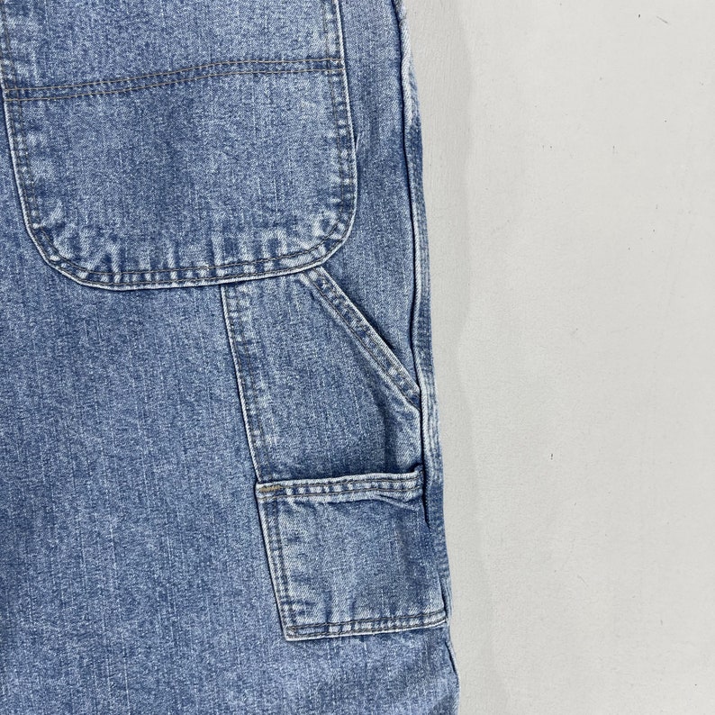 K&ouml;nnte beinhalten: Nahaufnahme einer blauen Jeans-Latzhose mit mehreren Taschen. Die Taschen sind mit einem kontrastierenden dunklen Faden vern&auml;ht.