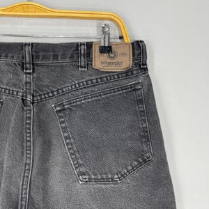 Pode incluir: Close-up da parte de tr&aacute;s de um par de jeans Wrangler pretos. Os jeans t&ecirc;m um design cl&aacute;ssico de cinco bolsos e um patch de couro com o logotipo Wrangler. Os jeans est&atilde;o pendurados em um cabide de metal.
