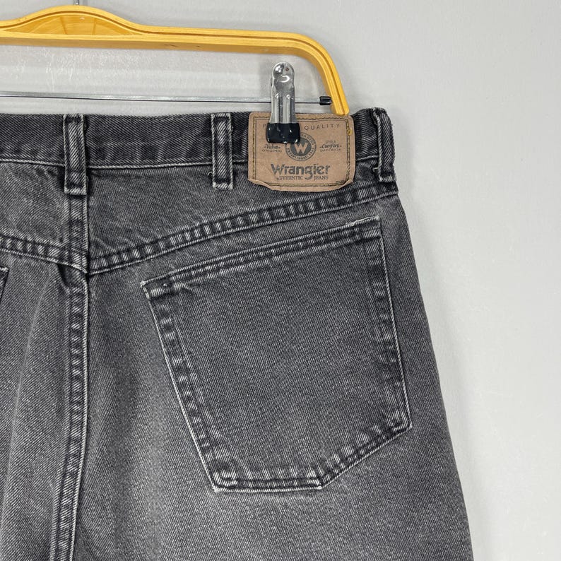 Pode incluir: Close-up da parte de tr&aacute;s de um par de jeans Wrangler pretos. Os jeans t&ecirc;m um design cl&aacute;ssico de cinco bolsos e um patch de couro com o logotipo Wrangler. Os jeans est&atilde;o pendurados em um cabide de metal.