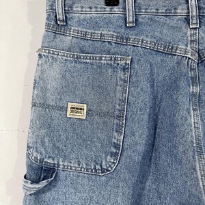 K&ouml;nnte beinhalten: Eine blaue Jeanshose im Carpenter-Stil mit einer gro&szlig;en Ges&auml;&szlig;tasche und einem Wrangler Originals-Patch. Die Jeans hat einen ausgewaschenen Look und ist aus strapazierf&auml;higem Material gefertigt.