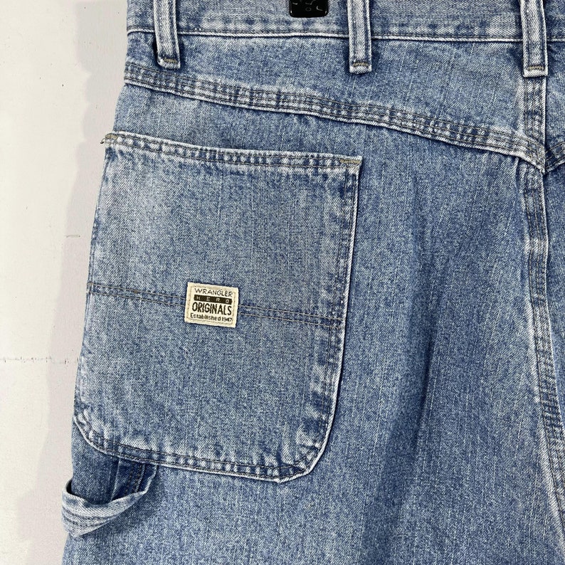 K&ouml;nnte beinhalten: Eine blaue Jeanshose im Carpenter-Stil mit einer gro&szlig;en Ges&auml;&szlig;tasche und einem Wrangler Originals-Patch. Die Jeans hat einen ausgewaschenen Look und ist aus strapazierf&auml;higem Material gefertigt.
