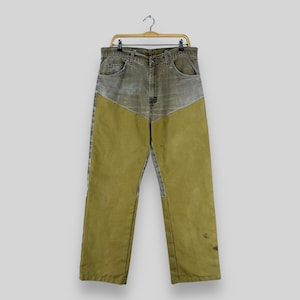 May include: Vintage denim jeans with a two-tone design. The upper portion is a faded gray-green, while the lower legs are a contrasting olive green. The jeans are hanging on a wooden hanger.