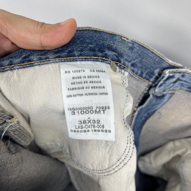 May include: Close-up of a pair of blue denim jeans with a white tag. The tag includes text indicating the jeans are made in Mexico, are 100% cotton, and have a 38x32 size. The jeans are slightly faded.