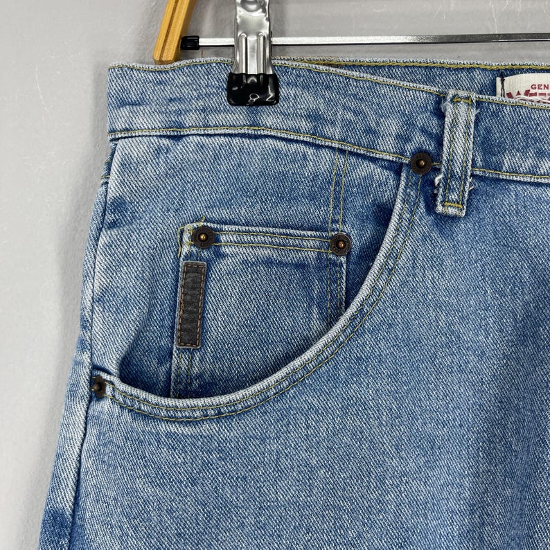 Puede incluir: Primer plano de unos vaqueros de mezclilla azul claro. Los vaqueros tienen un dise&ntilde;o cl&aacute;sico de cinco bolsillos con un bolsillo delantero visible. Las costuras son de un color contrastante y los vaqueros parecen desgastados.