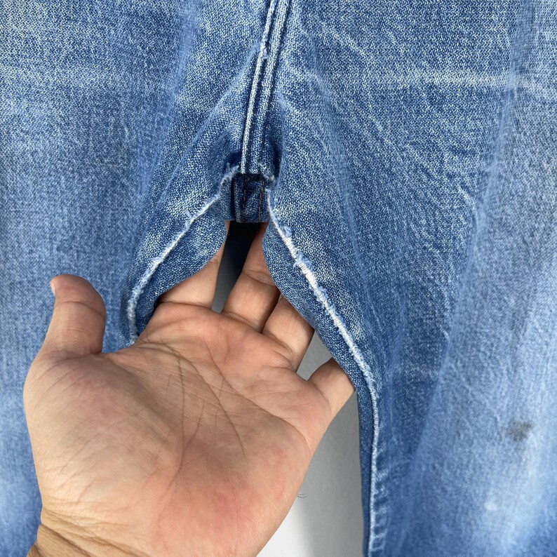 Pode incluir: Close-up de jeans de ganga azul claro. Os jeans mostram sinais de desgaste, com desbotamento vis&iacute;vel e um visual desgastado. Uma m&atilde;o segura os jeans abertos, mostrando o interior do bolso.