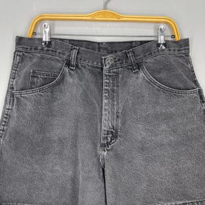 Pode incluir: Close-up de um par de cal&ccedil;&otilde;es de ganga cinzentos. Os cal&ccedil;&otilde;es t&ecirc;m um design cl&aacute;ssico de cinco bolsos, um fecho de bot&atilde;o e z&iacute;per e presilhas para cinto. Os cal&ccedil;&otilde;es est&atilde;o pendurados num cabide amarelo.
