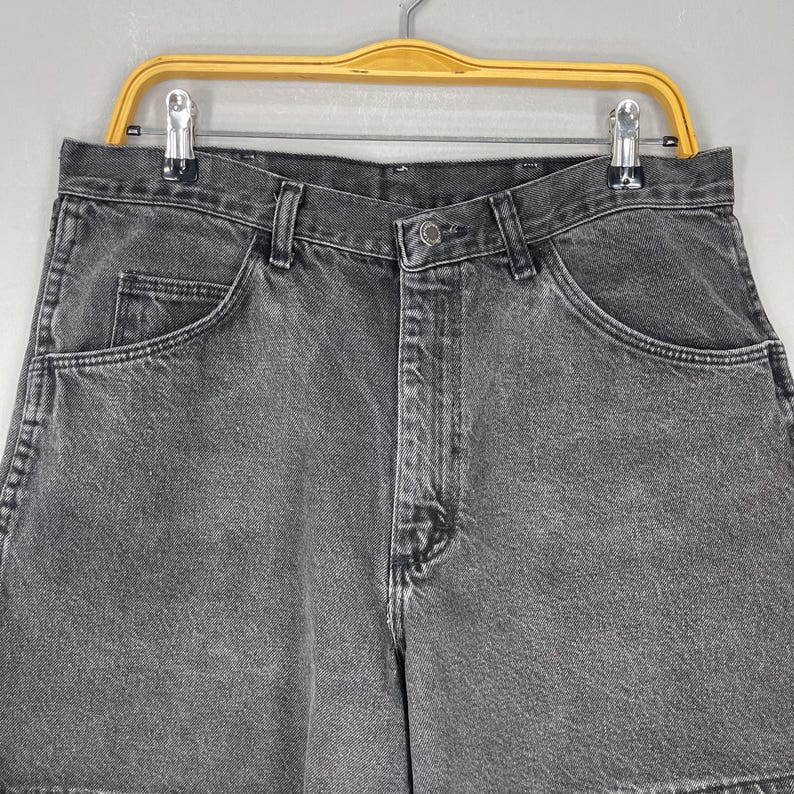 Pode incluir: Close-up de um par de cal&ccedil;&otilde;es de ganga cinzentos. Os cal&ccedil;&otilde;es t&ecirc;m um design cl&aacute;ssico de cinco bolsos, um fecho de bot&atilde;o e z&iacute;per e presilhas para cinto. Os cal&ccedil;&otilde;es est&atilde;o pendurados num cabide amarelo.