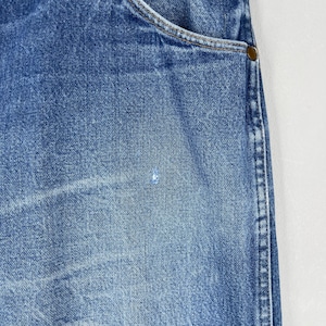 Pode incluir: Close-up de jeans de ganga azul. Os jeans mostram desgaste, com uma apar&ecirc;ncia desbotada e uma pequena mancha de cor clara. A borda do bolso e uma parte da costura s&atilde;o vis&iacute;veis.