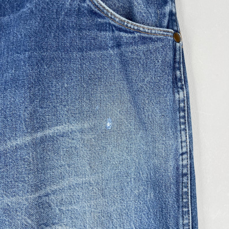 Pode incluir: Close-up de jeans de ganga azul. Os jeans mostram desgaste, com uma apar&ecirc;ncia desbotada e uma pequena mancha de cor clara. A borda do bolso e uma parte da costura s&atilde;o vis&iacute;veis.