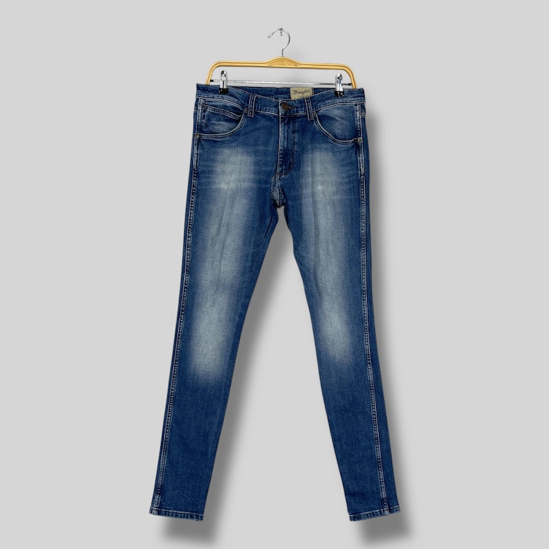 May include: Blue denim jeans hanging on a gold-colored hanger. The jeans have a classic five-pocket design, a button and zipper closure, and a faded wash. The jeans are straight-legged and appear to be new.