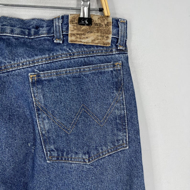 K&ouml;nnte beinhalten: Blaue Jeans aus Denim mit einer Ges&auml;&szlig;tasche mit einem markanten W-f&ouml;rmigen Steppdetail. Die Jeans haben einen braunen Leder-Patch mit dem Text "Wrangler" auf dem hinteren Bund.