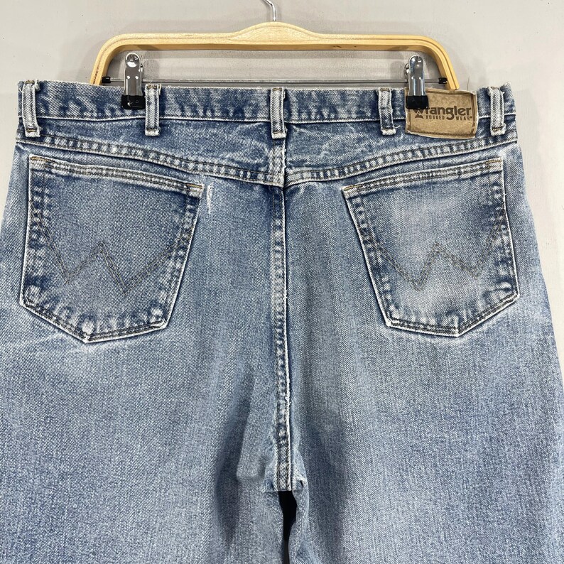 May include: A pair of light blue denim jeans with a Wrangler logo patch on the back right pocket. The jeans have a classic five-pocket style and a straight leg.